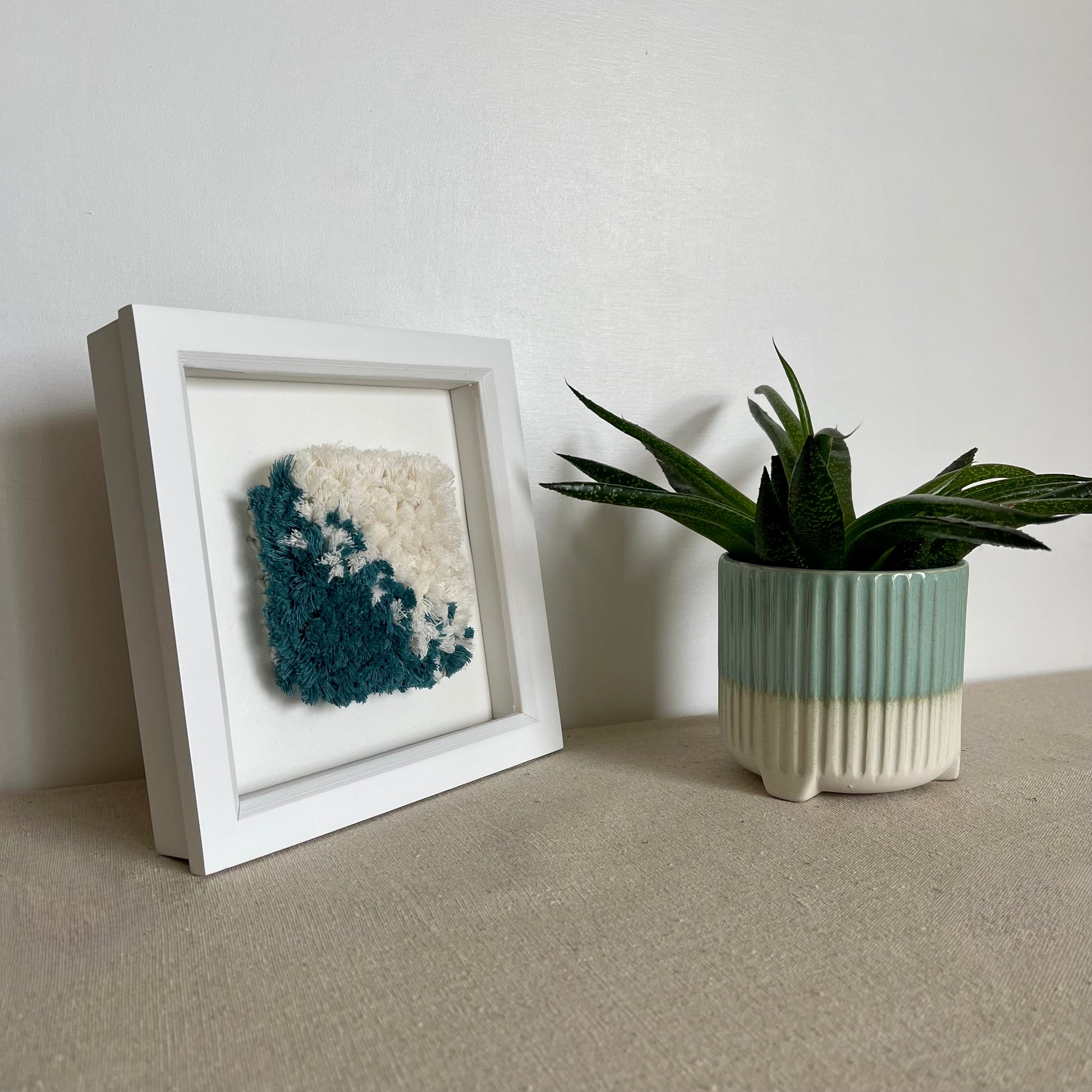Teal and cream fluffy textured woven textile art in a square white painted wood deep box frame next to plant