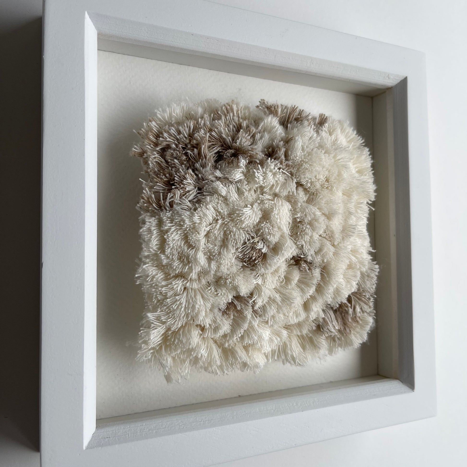 Angled view of cream and sand textured woven textile art in square white painted wood deep box frame on wall
