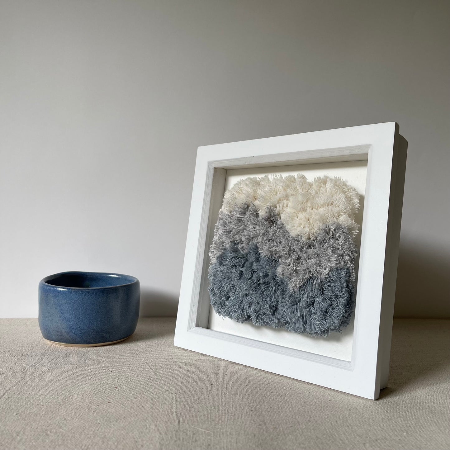 Cream and blue fluffy textured woven textile art in square white painted wood deep box frame next to blue pottery bowl