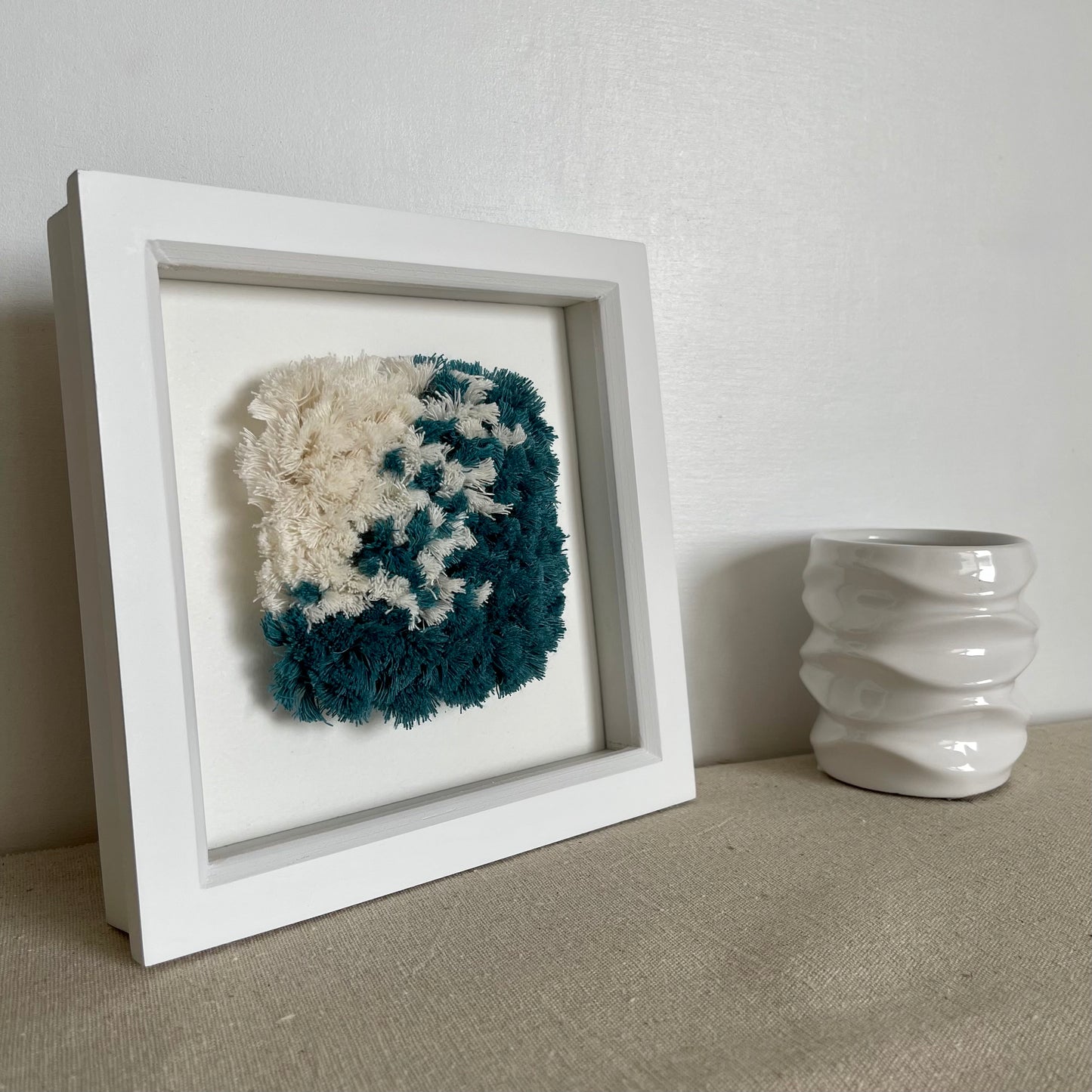Angled view of ocean teal and cream fluffy textured woven wall art in a square white painted wood frame