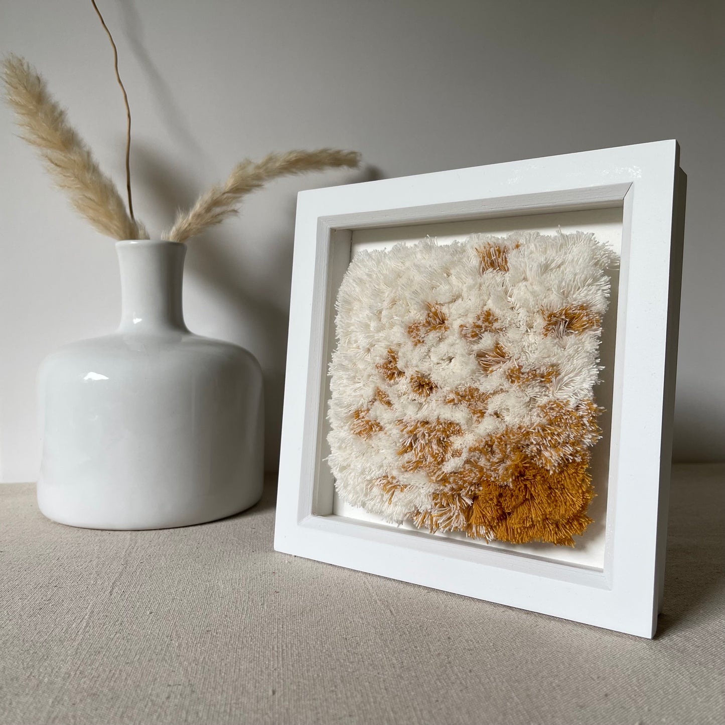 Cream and yellow ochre fluffy textured woven textile art in square white painted wood deep box frame on linen covered table next to scandi style vase