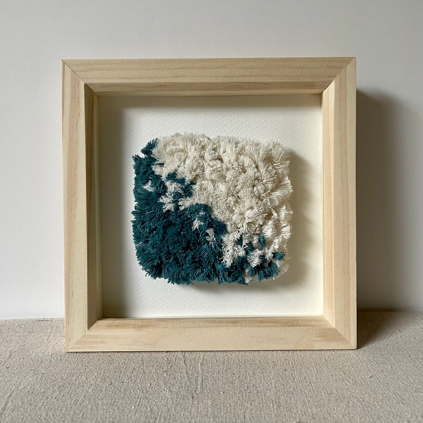 Front view of teal and cream hand woven textile art in a square natural wood deep box frame