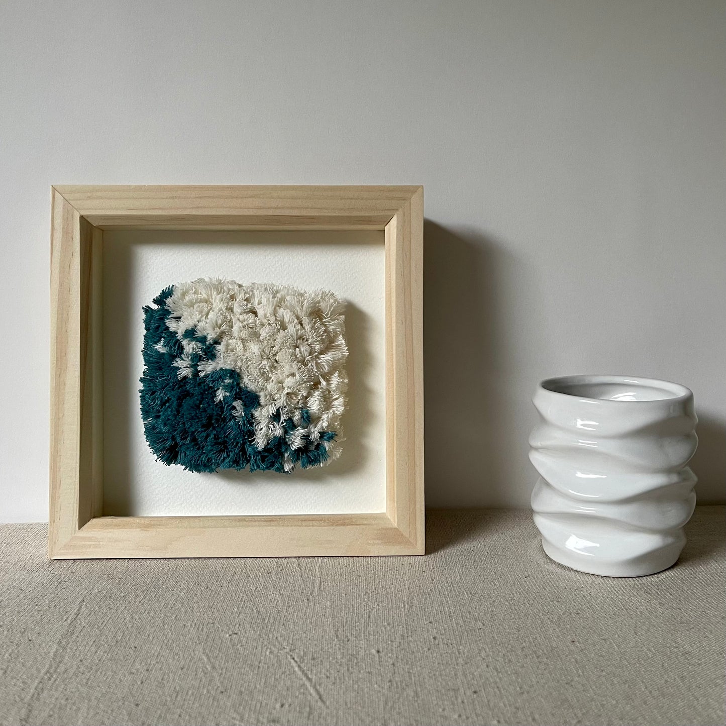 Teal and cream fluffy textured woven textile art in a square natural wood frame next to white ceramic vase
