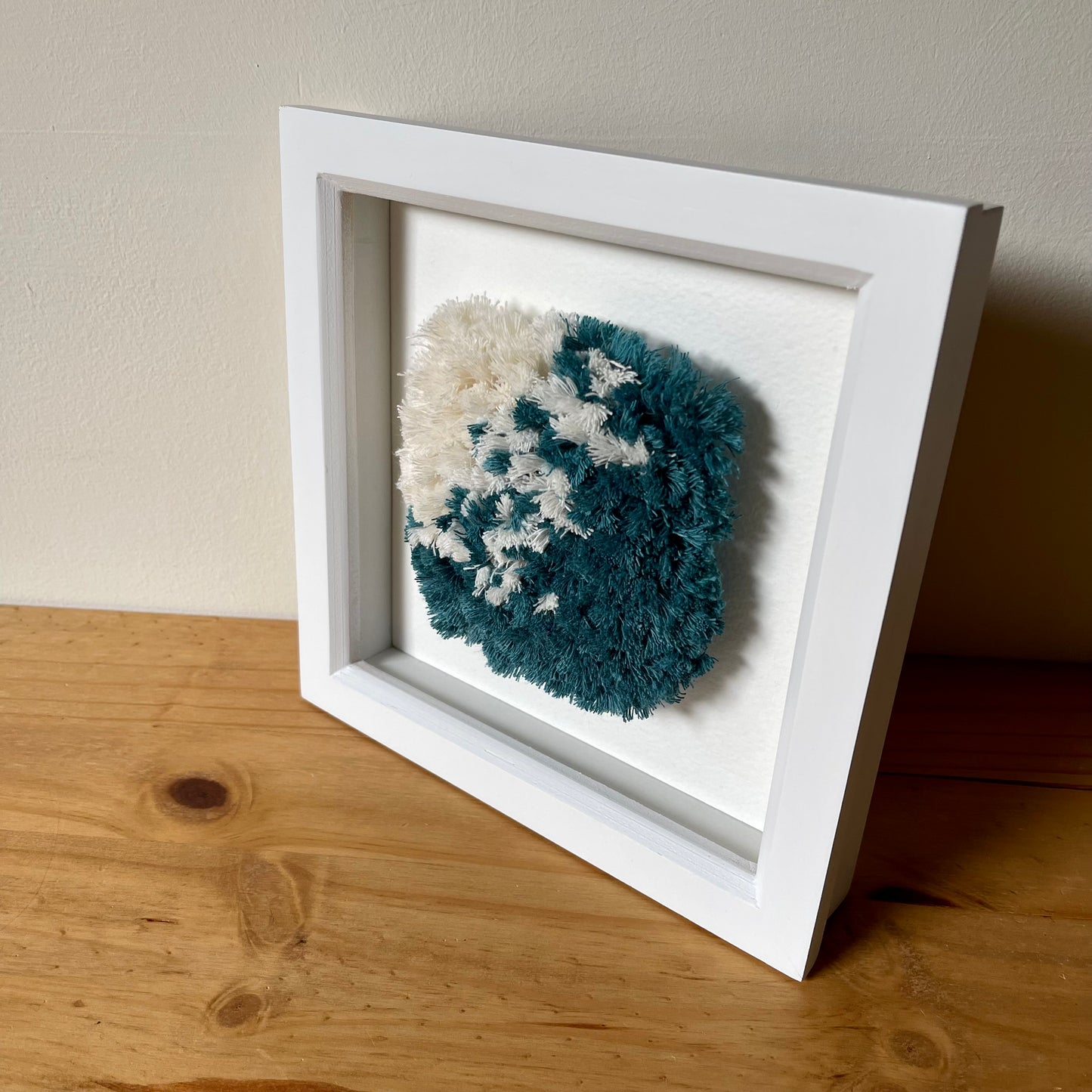 Square white painted wood deep box frame  containing a teal and cream fluffy textured woven piece of art