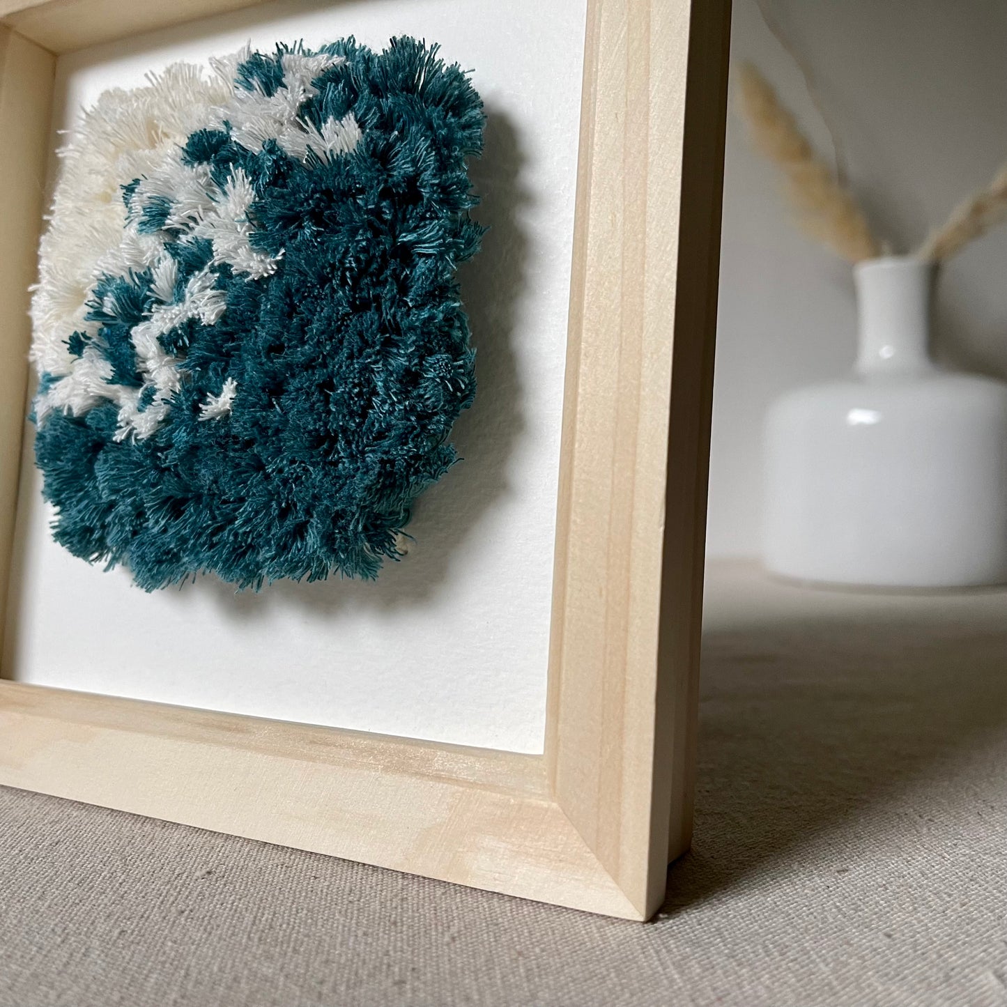 Angled close up view of ocean teal and cream fluffy textured woven art in a square wood box frame
