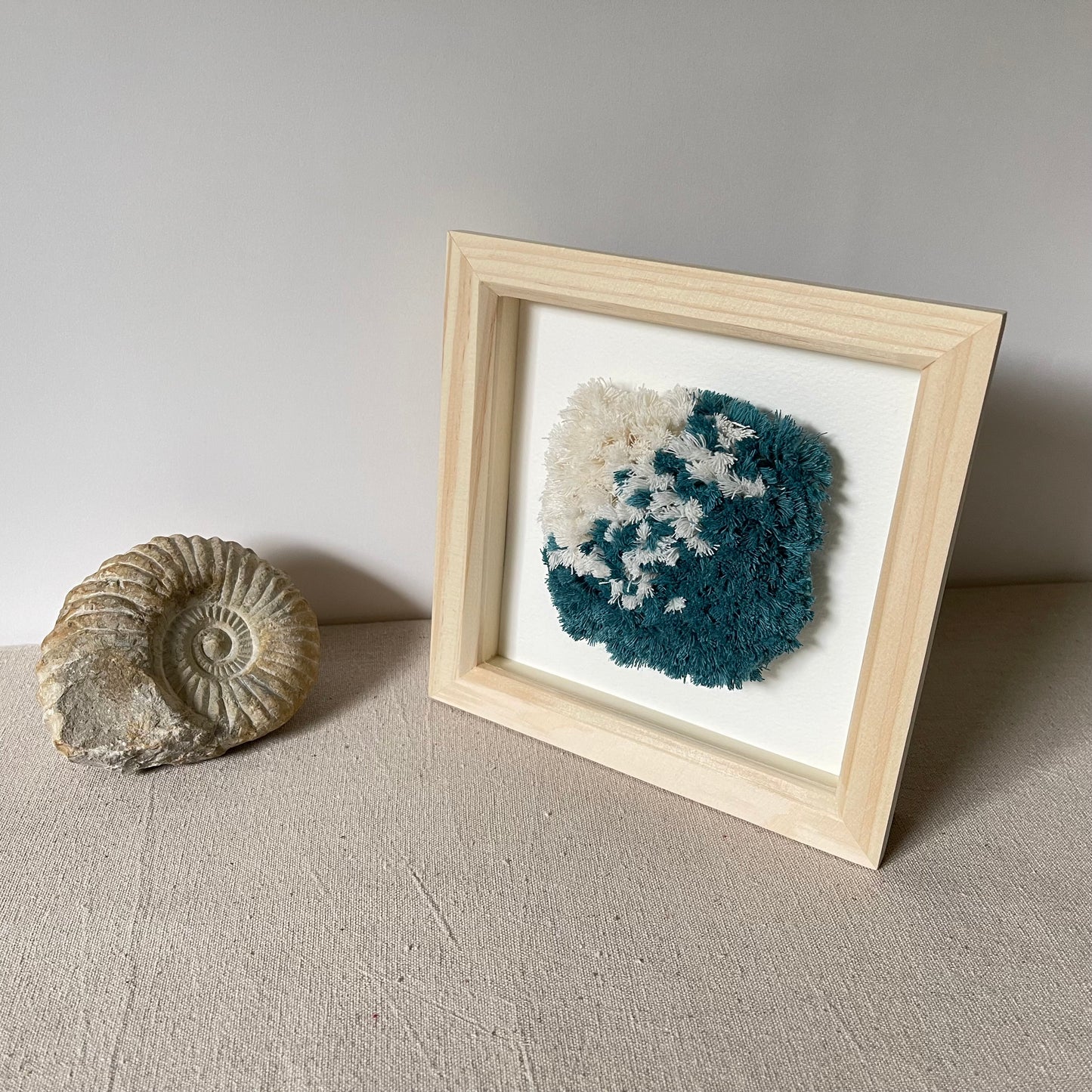 Ocean teal and cream fluffy textured woven art in a square wood deep box frame next to ammonite