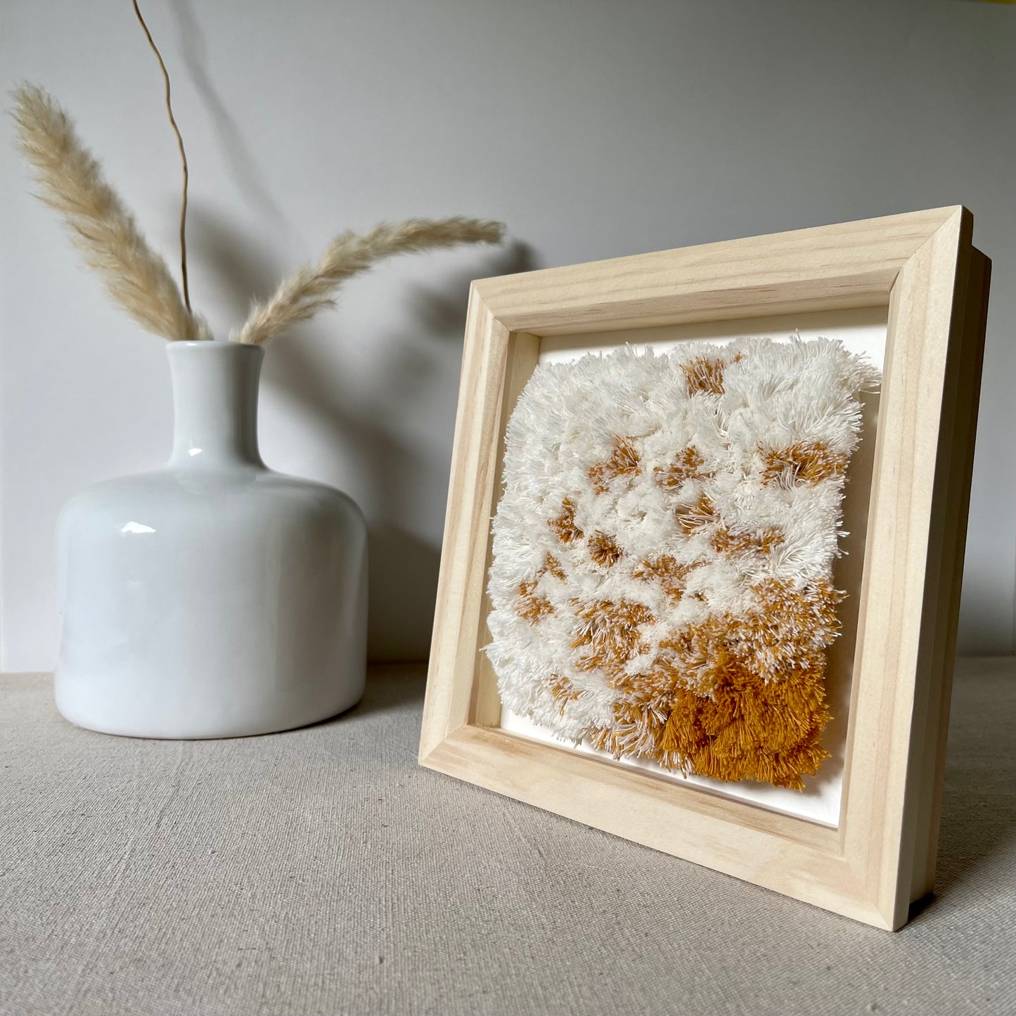 Cream and yellow ochre fluffy textured woven textile art in natural wood deep box frame on linen covered table next to scandi style vase