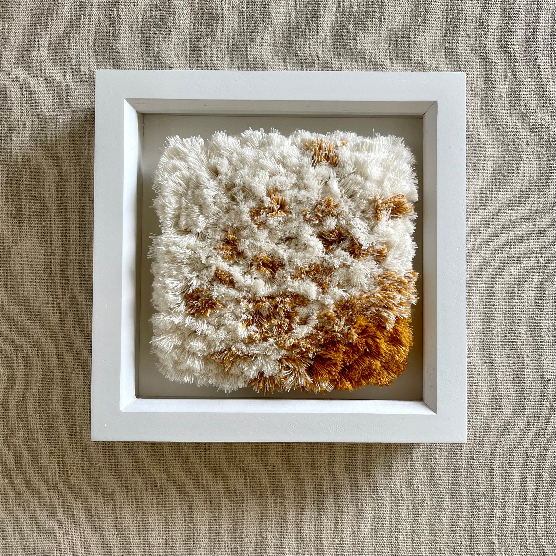 Front view of cream and yellow ochre fluffy textured woven textile art in square white painted wood deep box frame