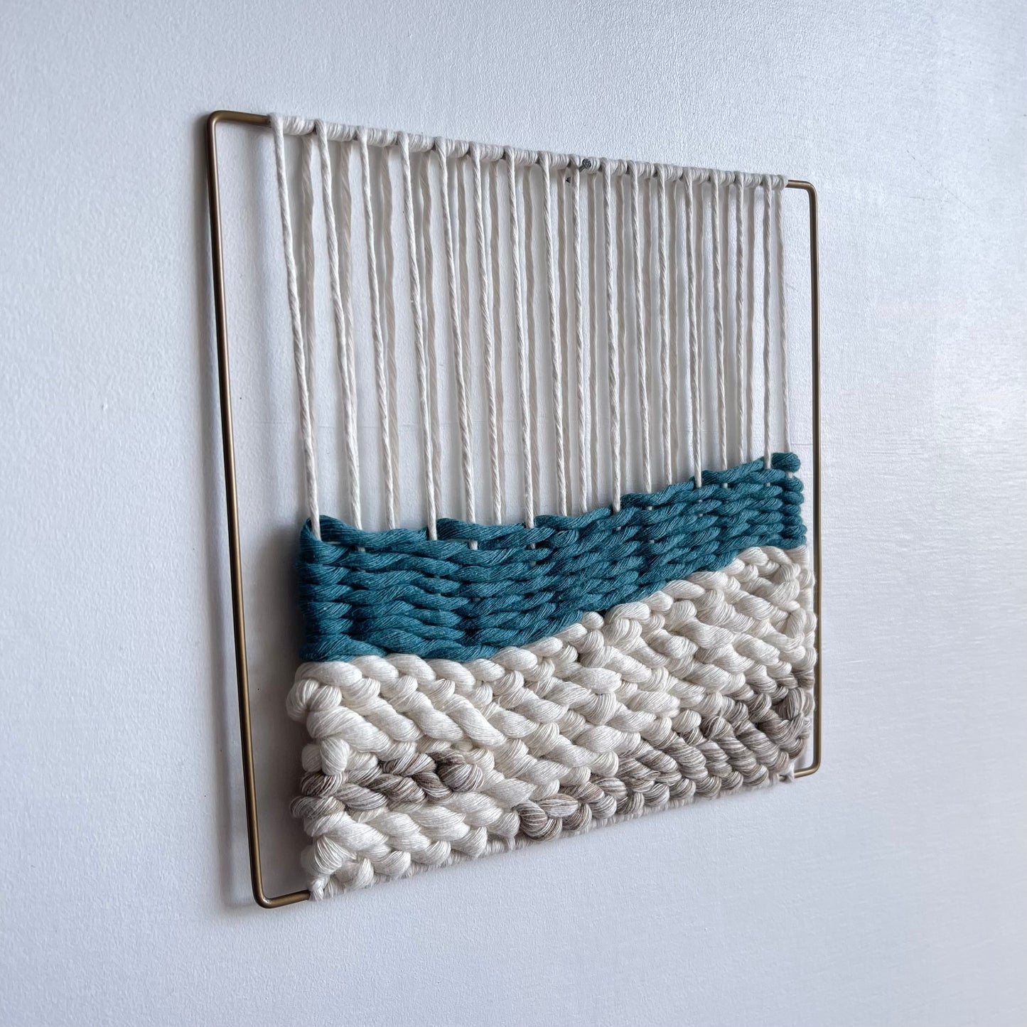 matt gold slim metal frame. Top section shows the warped thread around the frame that the weaving attaches to on the bottom half featuring a layer of teal over sand coloured cord simulating sea meeting sand.