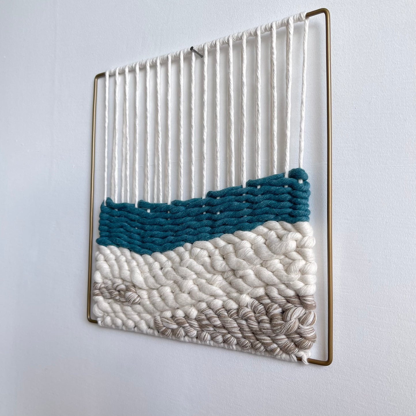 Square textured woven wall art on a matt gold slim metal frame. Top section shows the warped thread around the frame that the weaving attaches to on the bottom half featuring a layer of teal over sand coloured cord simulating sea meeting sand.