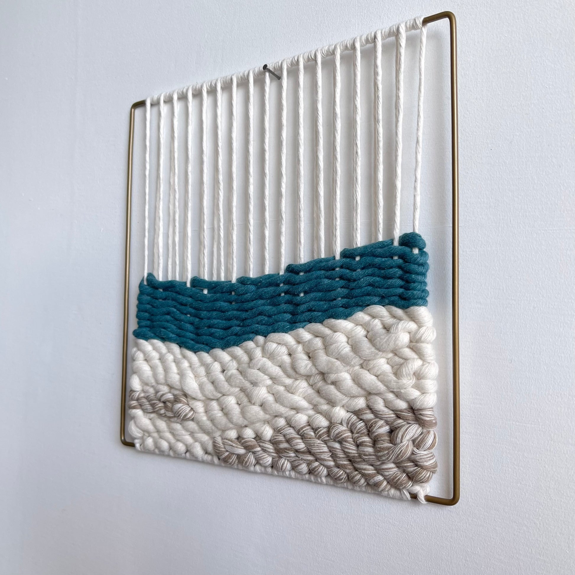 Square textured woven wall art on a matt gold slim metal frame. Top section shows the warped thread around the frame that the weaving attaches to on the bottom half featuring a layer of teal over sand coloured cord simulating sea meeting sand.