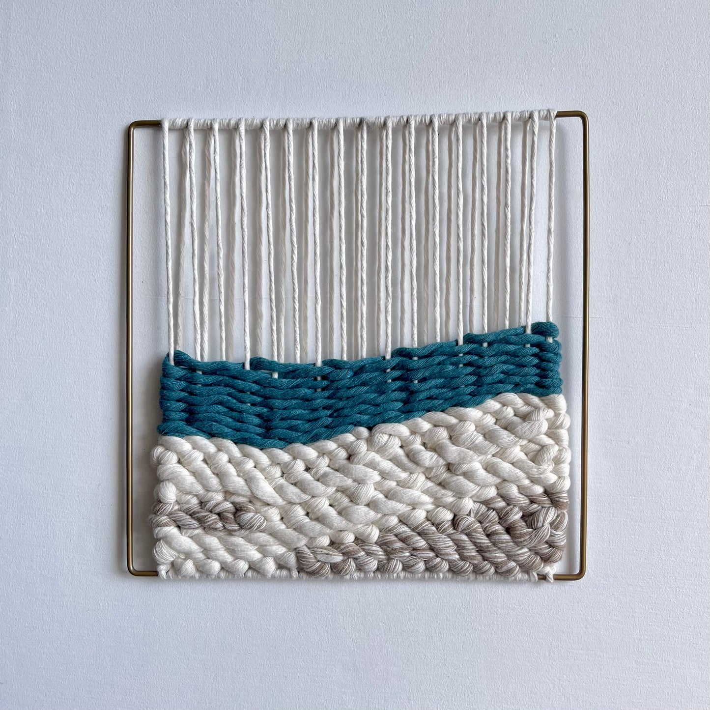 Square textured woven wall art on a matt gold slim metal frame. Top section shows the warped thread around the frame that the weaving attaches to on the bottom half featuring a layer of teal over sand coloured cord simulating sea